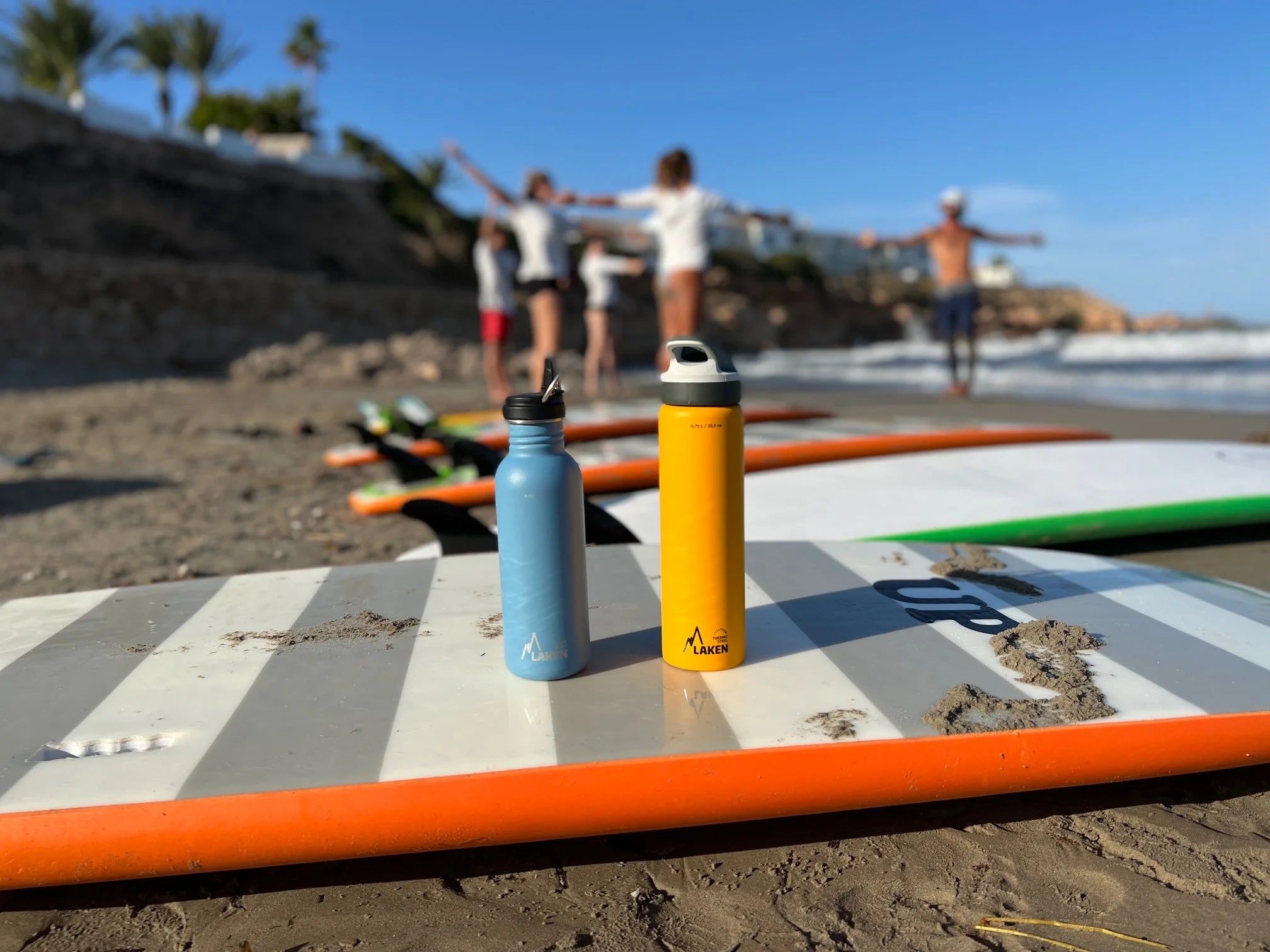 Una botella térmica LAKEN y otra de acero encima de una tabla de surf en la playa