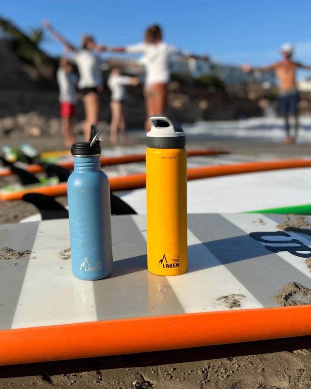 Una botella térmica LAKEN y otra de acero encima de una tabla de surf en la playa